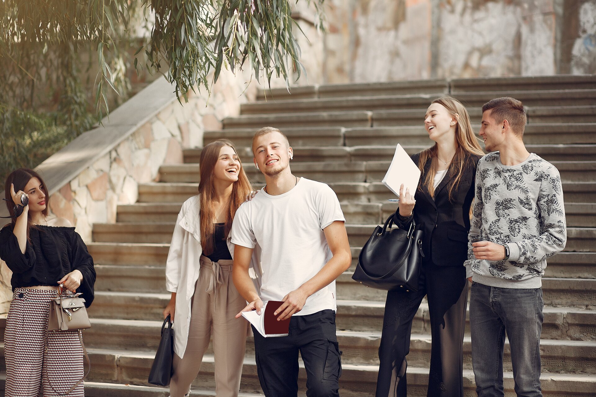 Ilustracja przedstawia grupę młodych ludzi schodzących ze schodów. Po lewej stronie ilustracji znajduje się kobieta ubrana w czarną koszulkę oraz jasne spodnie. Na środku ilustracji znajduje się mężczyzna i kobieta. Mężczyzna ubrany jest w białą koszulkę oraz czarne spodnie, kobieta zaś w jasną koszulkę i jasne spodnie. Po prawej stronie ilustracji stoi kolejna para. Mężczyzna ubrany jest w szarą bluzę oraz czarne spodnie, kobieta w czarny kombinezon. Na drugim planie znajdują się jasne schody. 