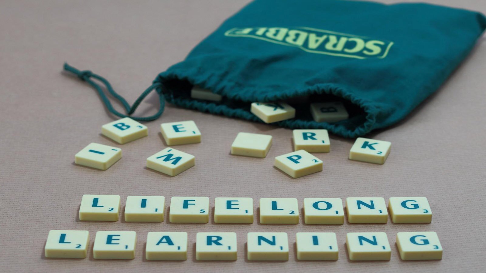 Zdjęcie przedstawia leżący na stole zielony woreczek popularnej gry w scrabble. Z rozsypanych kostek z literkami ułożono napis w języku angielskim:  LIFELONG LEARNING.