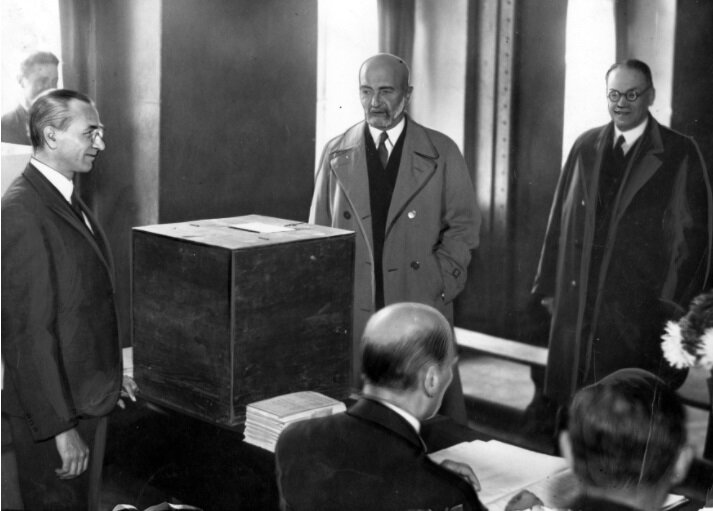 Zdjęcie przedstawia wnętrze pomieszczenia. W części głównej widać urnę wyborczą, wokół której stoi kilku mężczyzn. Inni mężczyźni siedzą za stołem, na którym leżą ułożone w stos papiery.