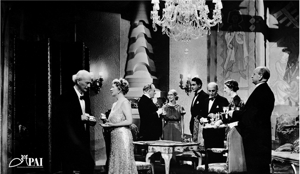 Fotografia przedstawia Ignacego Jana Paderewskiego i Helenę Modrzejewską, którzy rozmawiają ze sobą stojąc w salonie, w otoczeniu innych osób - uczestników przyjęcia. W dłoniach trzymają filiżanki, są elegancko ubrani.