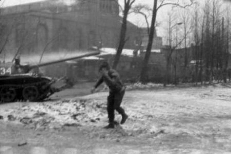 Zdjęcie przedstawia plac, po którym biegnie mężczyzna. Na wprost niego jedzie czołg. W tle budynek kopalni i drzewa.