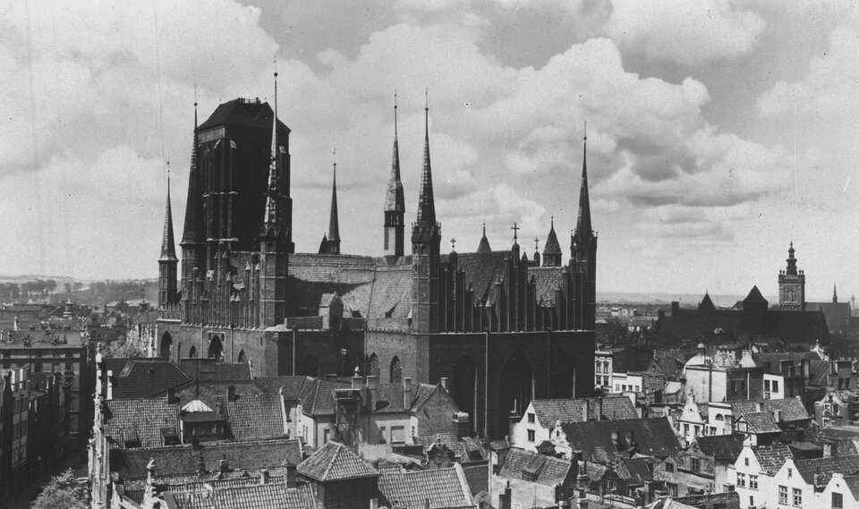 Zdjęcie przedstawia śródmieście gdańska. Na tle gęsto zabudowanych kamienic wyróżnia się budynek gotyckiej świątyni ze strzelistymi wieżyczkami oraz wysoką wieżą po lewej stronie. Na ścianach widoczne są wysokie okna zakończone łukami. W tle zdjęcia jest niebo z chmurami.