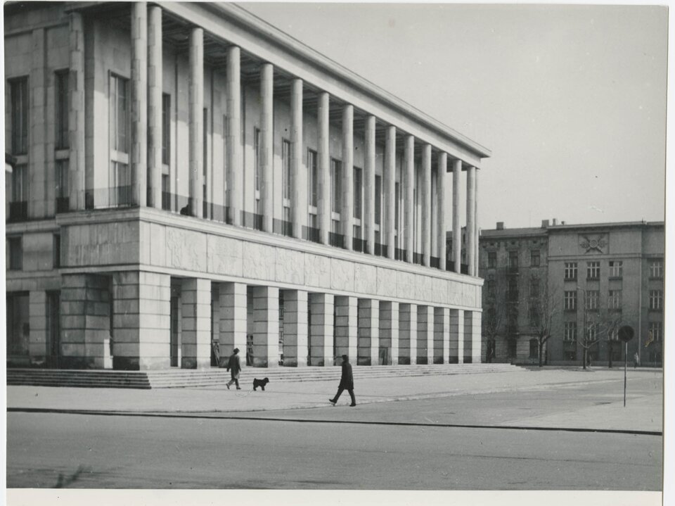 Zdjęcie przedstawia kilkupiętrowy budynek socrealistyczny, który stoi przy wybetonowanym placu. Jego elewacja jest wykonana z kamiennych płyt. Budynek przy podstawie składa się z grubych kolumn, nad nimi jest frontowa elewacja z płaskorzeźbami. Wyżej są wysokie okna, przed którymi jest rząd smukłych kolumn.