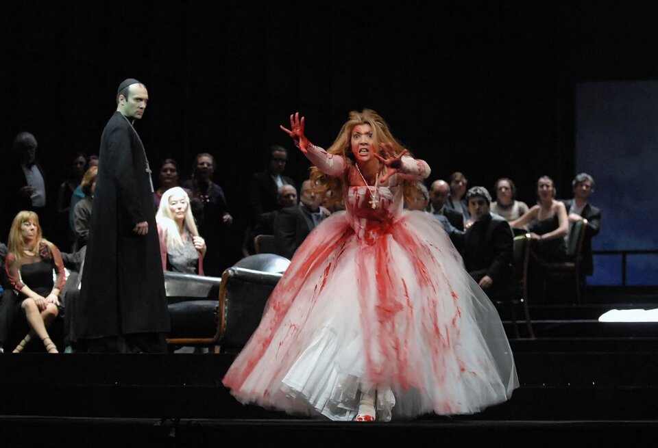 Ilustracja przedstawia scenę obłędu z opery G. Donizettiego Łucja z Lammermoore. Kobieta w białej, długiej, okrwawionej sukni. Stoi, ma ręce odchylone do przodu. Na szyi ma łańcuch ze srebrnym, dużym krzyżem. Kobieta ma długie, rude włosy. Obok kobiety znajduje się ksiądz w czarnym stroju.