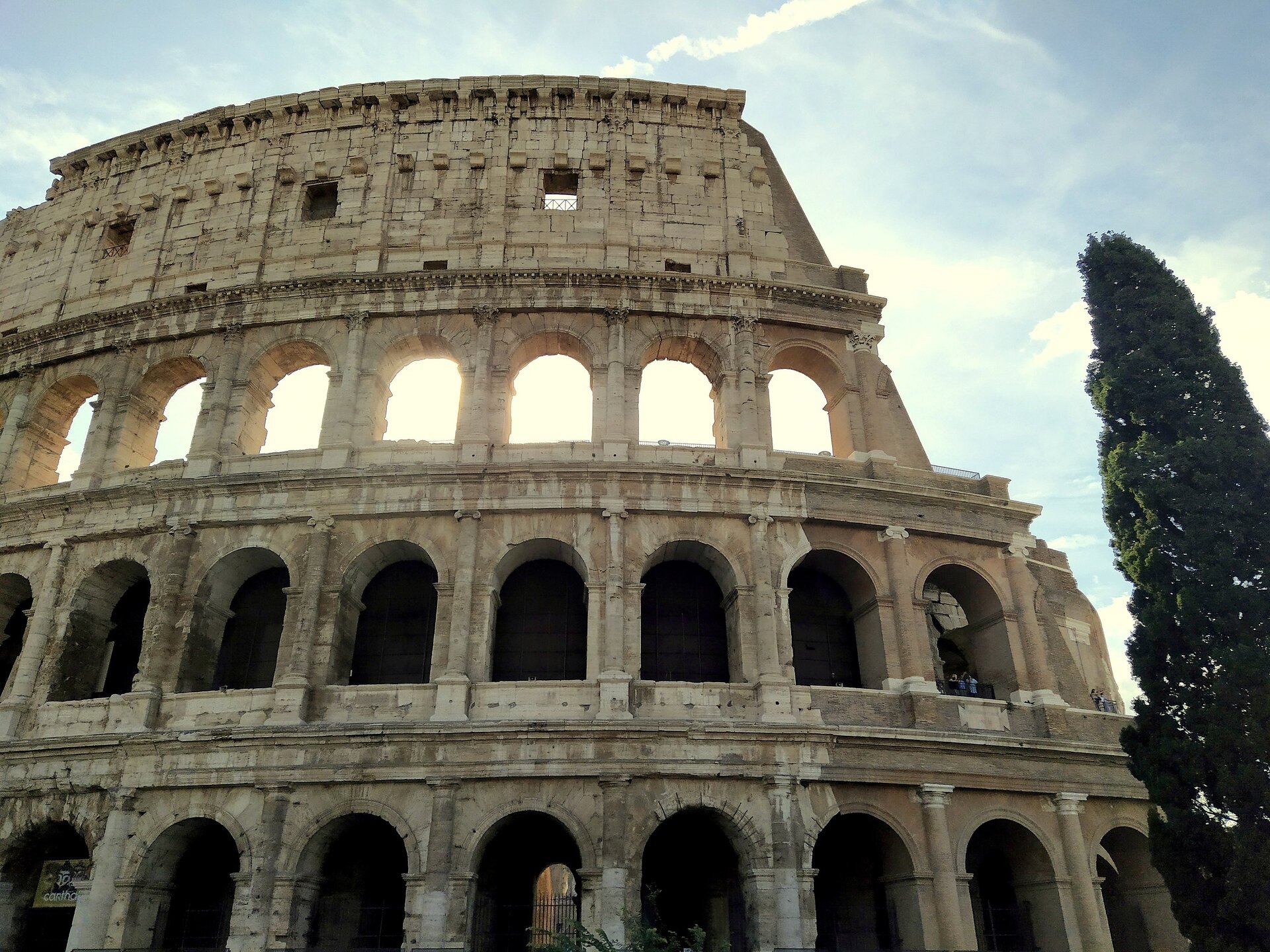 Zdjęcie przedstawia fragment monumentalnej budowli zbudowanej na planie elipsy. Budowla jest częściowo zrujnowana, nie ma prawej części. Składa się ona z ustawionych na sobie na czterech poziomach szeregu łuków architektonicznych. Czwarty poziom, najwyższy i ostatni, nie ma struktury łuków, jest zabudowany i pokryty niewielkimi zdobieniami. Przez łuki na trzecim poziomie przebija światło. Budowla ma kolor żółto‑piaskowy. Po prawej stronie budowli widać fragment wysokiego drzewa przypominającego cis. Niebo jest jasne i raczej bezchmurne.