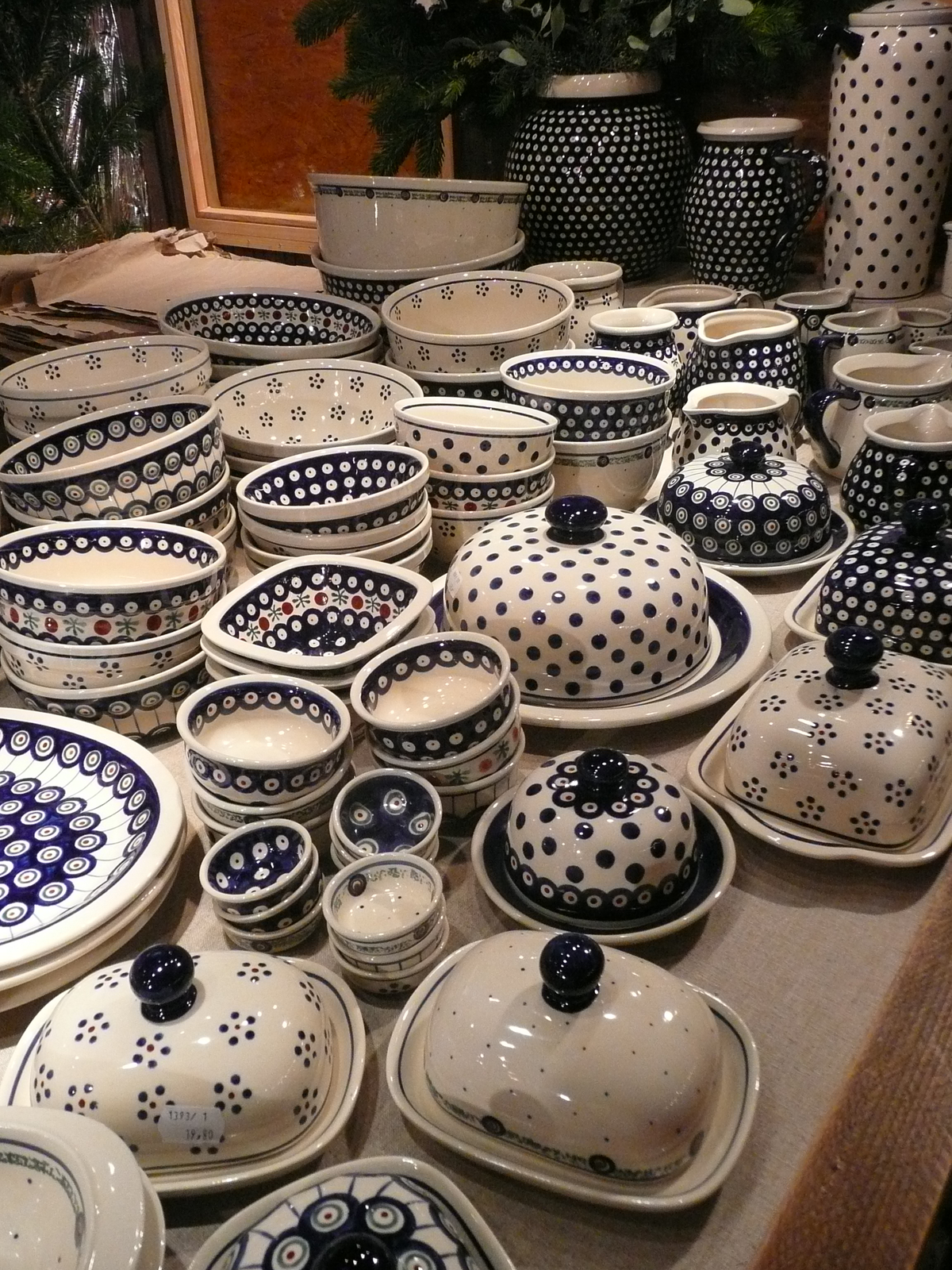 Zdjęcie przedstawiające ceramikę Bolesławiec. Liczne misy i miseczki, a także wazony i maselniczki z charakterystycznymi biało-granatowymi wzorami, nawiązującymi do pawich oczek, jagód, czy też kwatów.