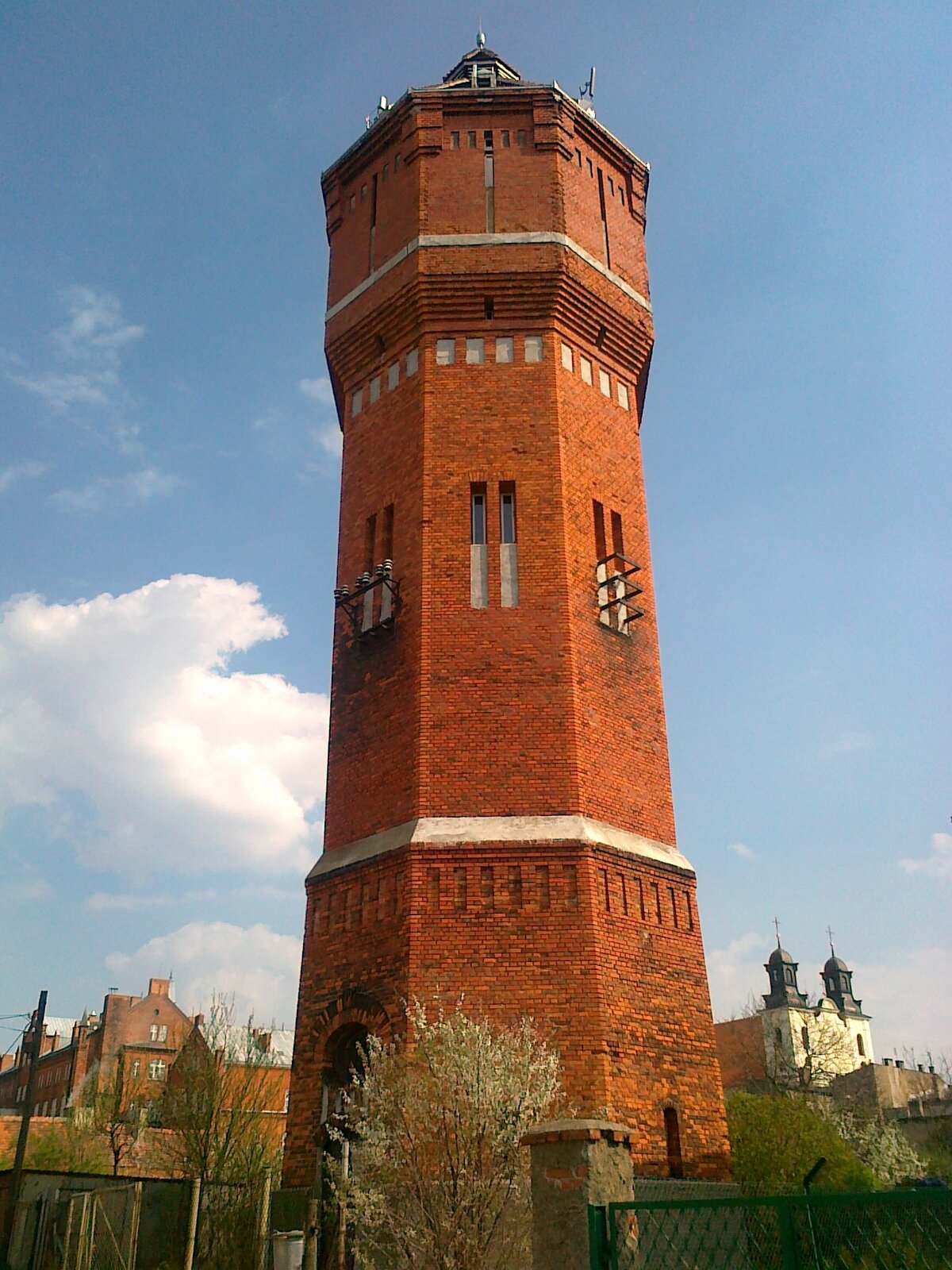 Kliknij, aby powiększyć Zdjęcie przedstawia ceglaną wieżę zbudowaną na planie heksagonu. Posiada wąskie, wysokie okna, zlokalizowane w jej górnej części (na poddaszu i piętrze niżej). Za wieżą znajdują się zabudowania miasta - domy i kościół. W tle niebo.
