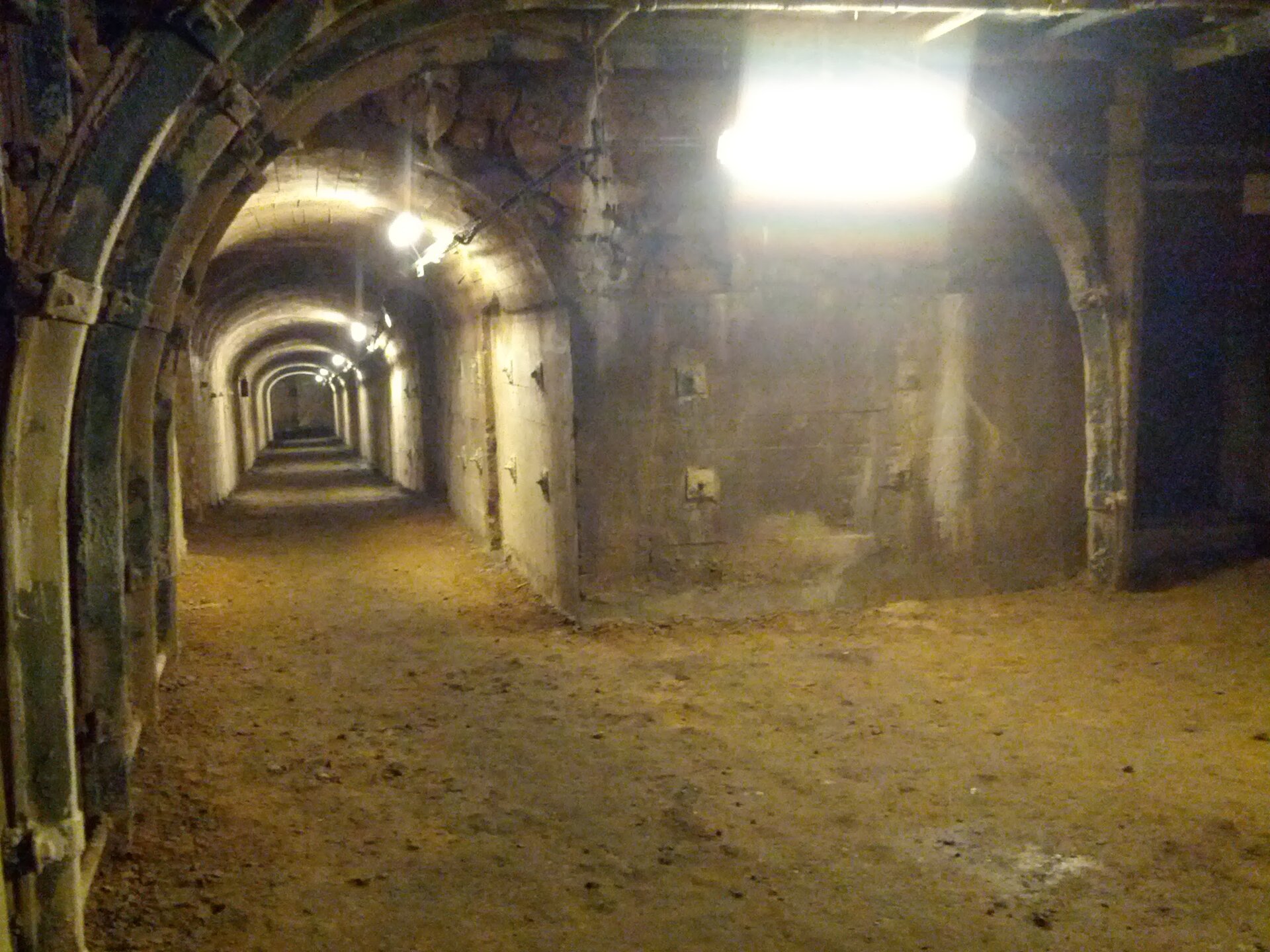 Fotografia przedstawia tunel lub piwnicę. Ściany tunelu zbudowane są z kamiennych płyt, a podłoga pokryta jest piachem. Na ścianie wisi oświetlenie elektryczne.