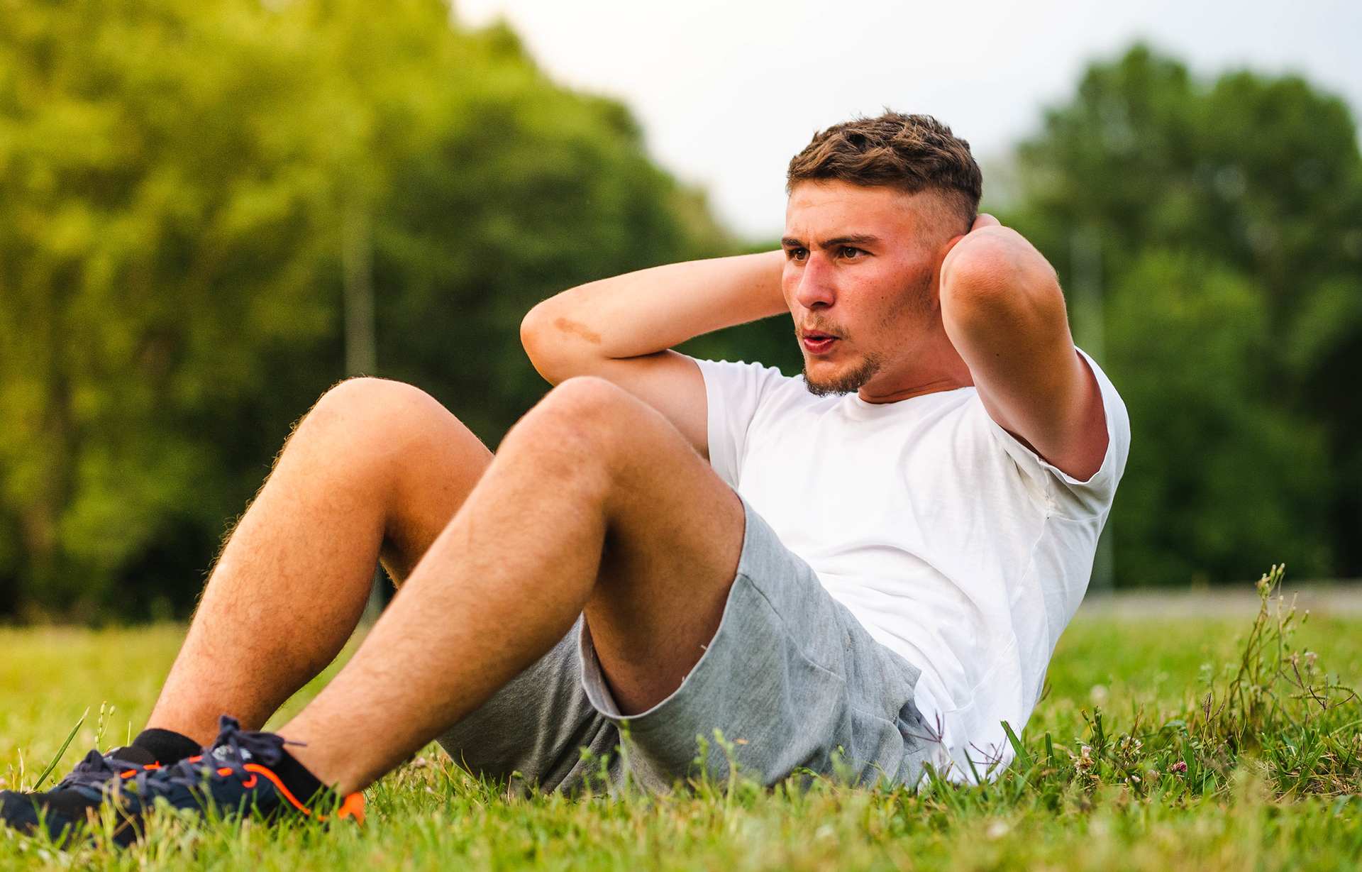 Zdjęcie przedstawia młodego mężczyznę ćwiczącego brzuszki na trawie. Mężczyzna ma krótkie, ciemne włosy, jest ubrany w białą koszulkę z krótkim rękawem, szare szorty i czarne, sportowe buty. Siedzi na trawie, ma podciągnięte do góry kolana, obie ręce założone na kark. W tle widoczne są rozmyte drzewa.