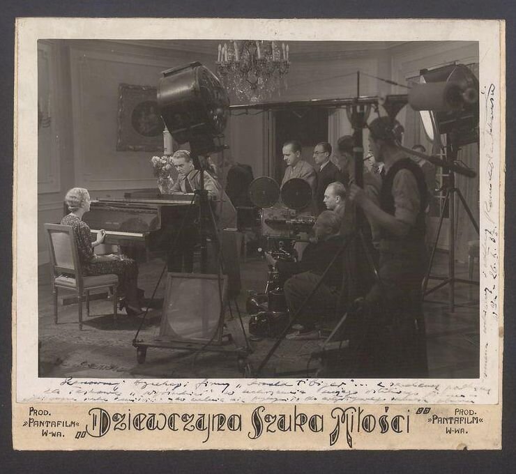 Zdjęcie przedstawia plan filmu. Po lewej stronie aktorka siedzi przy fortepianie. Po prawej stronie ustawiony jest sprzęt: kamery, oświetlenie oraz mikrofony. Sprzęt jest obsługiwany przez grupę mężczyzn.