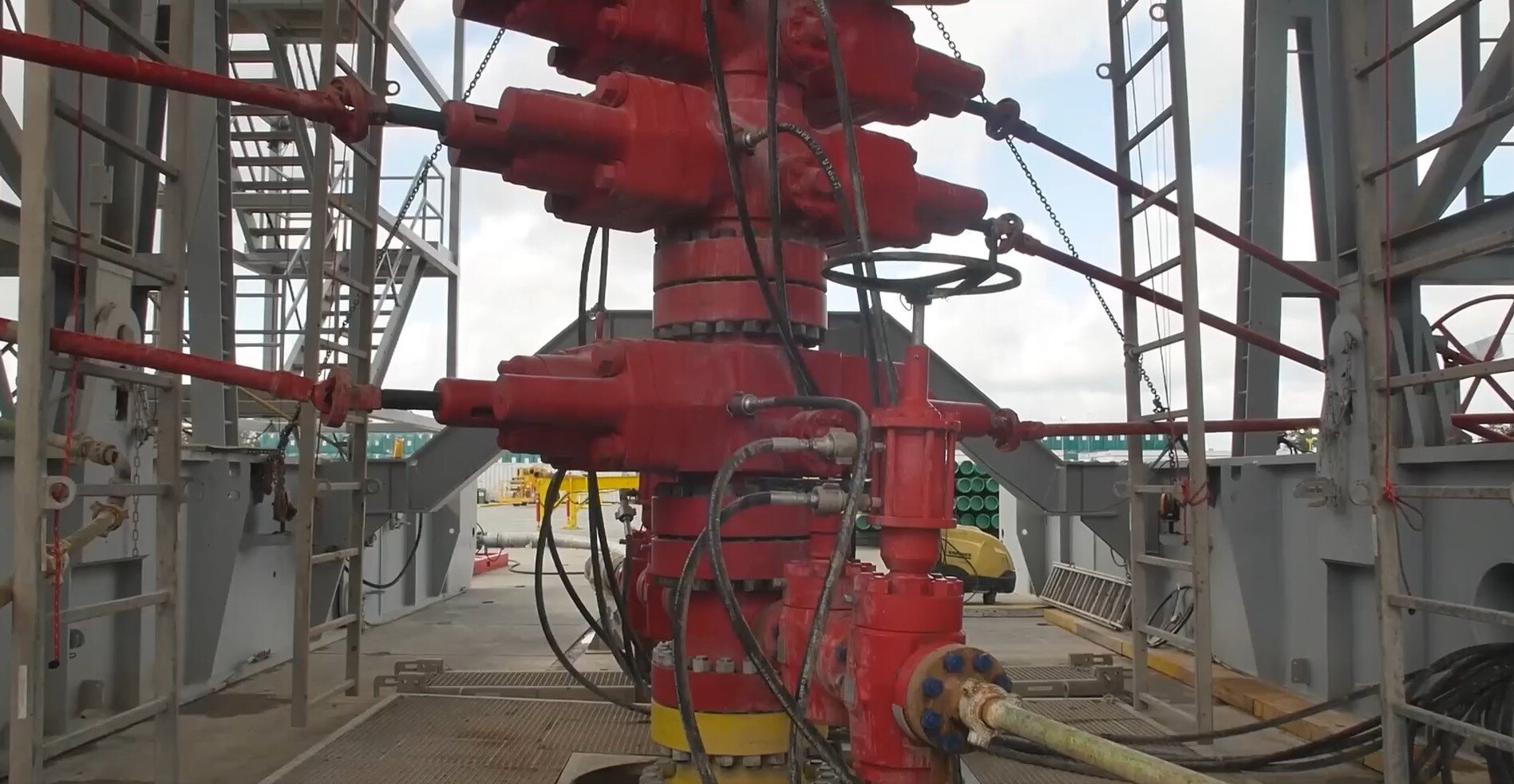  Zdjęcie przedstawia czerwone stalowe uzbrojenie przeciwerupcyjne otworu. Elementy w kształcie walca połączone są u podstawy oraz na wysokości środka urządzenia łącznikiem kołnierzowym połączonym przy użyciu czarny ch śrub. Z boku urządzenia, na wysokości podstawy, środka urządzenia oraz szczytu urządzenia, odchodzą czerwone liny .