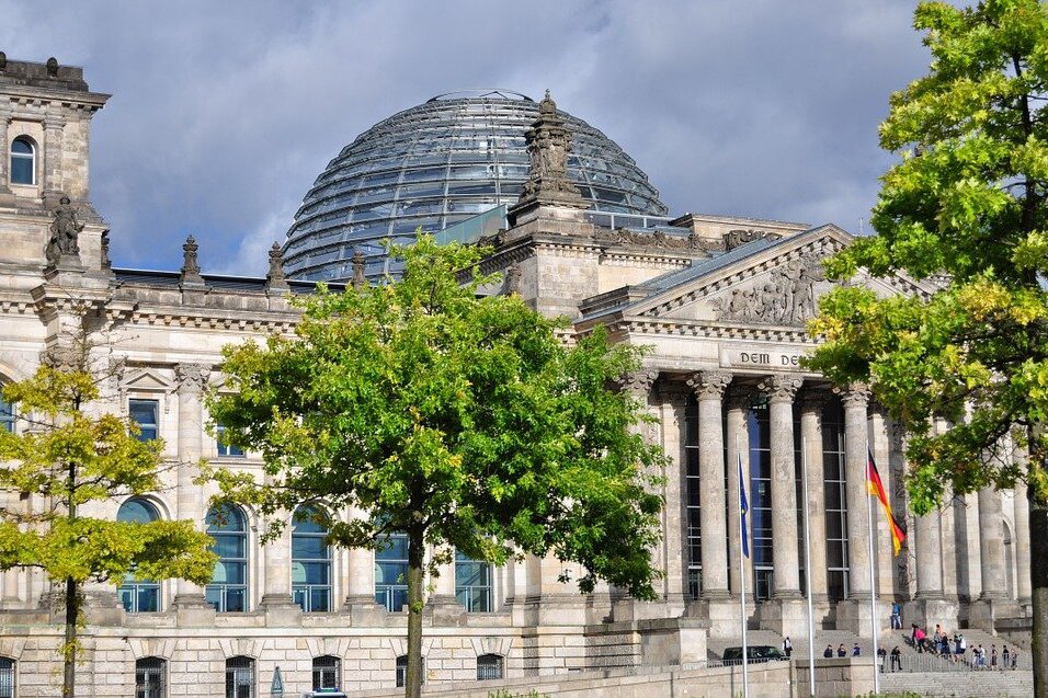 Zdjęcie przedstawia Gmach Parlamentu w Berlinie. Ma charakterystyczną szklaną kopułę. Widać ludzi wchodzących do budynku oraz flagę Niemiec na wysokim maszcie przed wejściem. Do budynku prowadzą szerokie betonowe schody.