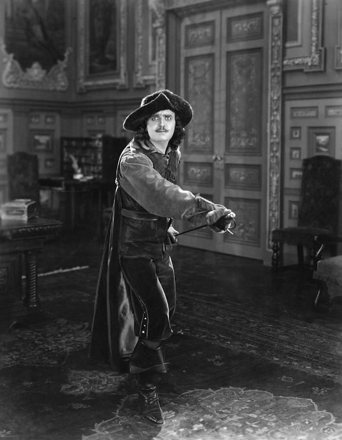 Kliknij, aby powiększyć. Zdjęcie przedstawia mężczyznę, stojącym na tle bogato zdobionej komnaty. Ma on dłuższe, sięgające do ramion, kręcone włosy oraz czarnego wąsa. Na jego twarzy widać zaskoczenie i przerażenie. Na głowie ma czarny kapelusz muszkietera. Ma on jasną koszulę z bufiastymi rękawami, ciemną kamizelkę i ciemne, luźne spodnie. Do kamizelki ma przypięty czarny, długi płaszcz. Mężczyzna prawą dłonią próbuje wysunąć szpadę, która przymocowana jest do jego lewego biodra. Zdjęcie jest czarno‑białe.