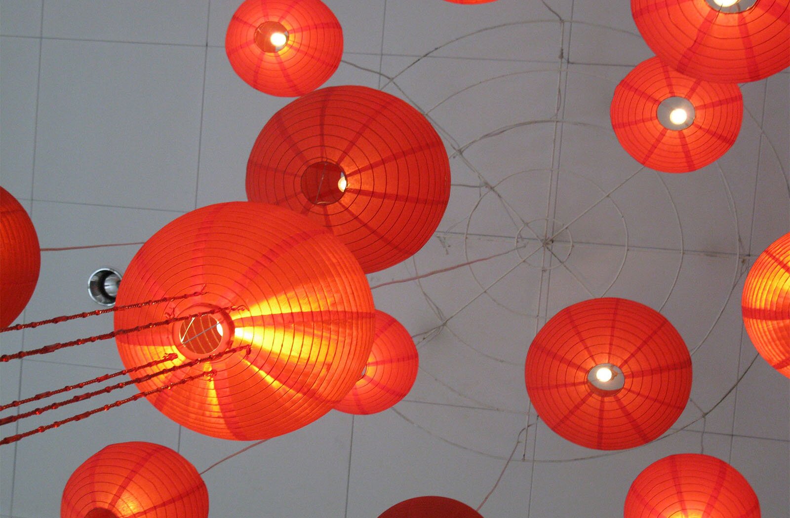 Zdjęcie przedstawia czerwone, papierowe lampiony. Lampiony są zawieszone na sznureczkach i podwieszone na konstrukcji przypominającej pajęczynę.