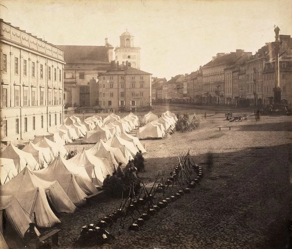 Zdjęcie przedstawia duży plac, na którym znajdują się stojące szeregami namioty wojskowe. Przed namiotami siedzą żołnierze w mundurach, z karabinami. Niedaleko nich leżą hełmy i stoją ustawione karabiny.