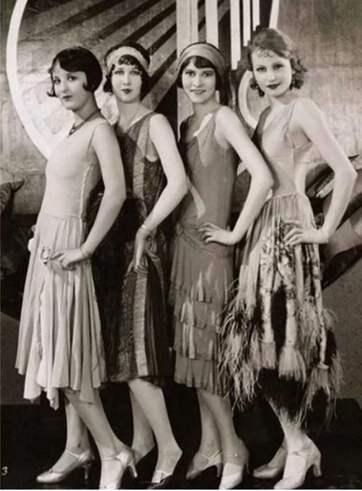 Zdjęcie przedstawia cztery szczupłe kobiety, które stoją bokiem z rękoma na ramionach. Ubrane są w sukienki na ramiączkach, które sięgają łydek. Mają włosy o długości do ucha. Dwie z nich mają na włosach opaski.