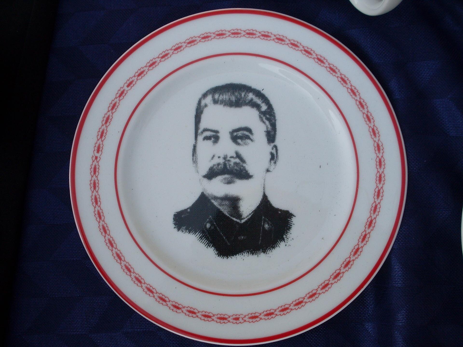 Zdjęcie przedstawia biały talerz z czarnym wizerunkiem Józefa Stalina. Widoczne jest jego popiersie. Ma gęste wąsy, zaczesane do tyłu włosy oraz ubrany jest w mundur. Talerz posiada czerwone obramowanie z prostym wzorem.