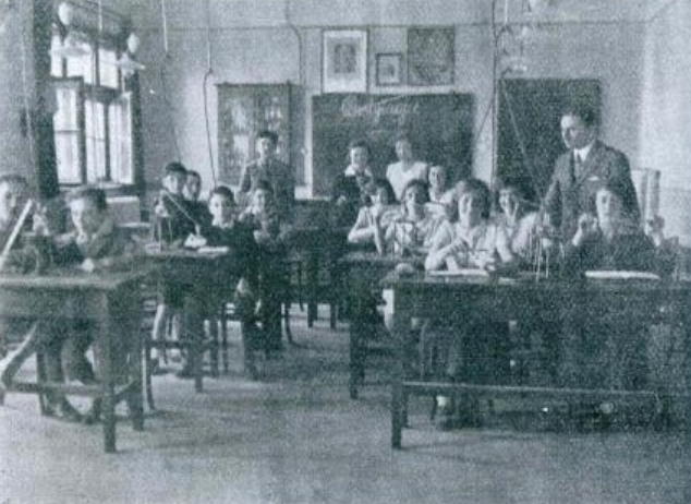 Fotografia przedstawia klasę w jednej ze szkół. Uczniowie siedzą w ławkach i patrzą w obiektyw. Między ławkami przechadza się nauczyciel. Widać tablicę i wiszące nad nią obrazy. Uczniowie mają na ławkach pomoce naukowe.