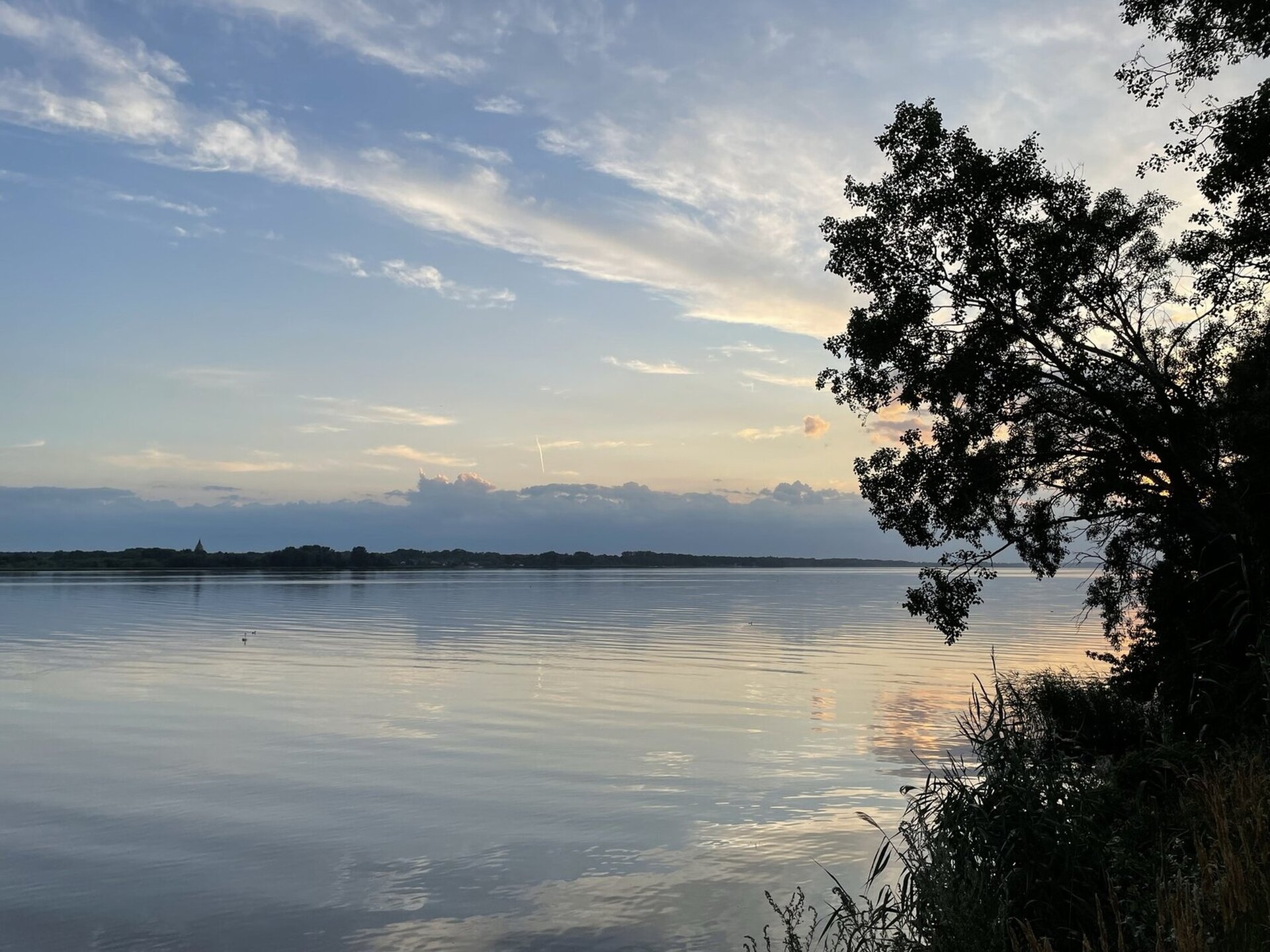 Fotografia przedstawia szeroką rzekę. Po prawej stronie fragment brzegu, na którym są drzewa. W oddali drugi brzeg. Na niebie kłębiaste chmury, przysłaniające słońce. 