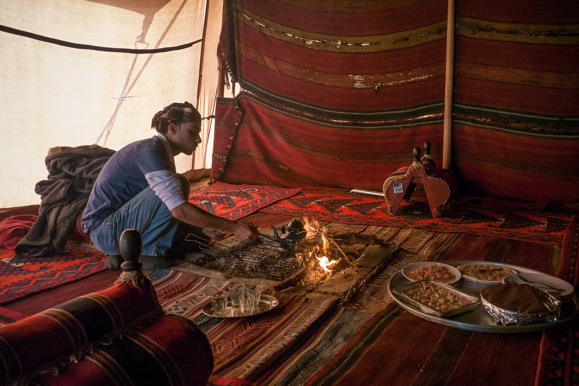 Zdjęcie przedstawia wnętrze namiotu. Okryte jest kolorowym materiałem. Na środku palenisko. Obok taca z jedzeniem. Mężczyzna poprawia palenisko, obok na talerzach i tacy przygotowane jedzenie do przyrządzenia. Wokół paleniska rozłożone dywany.