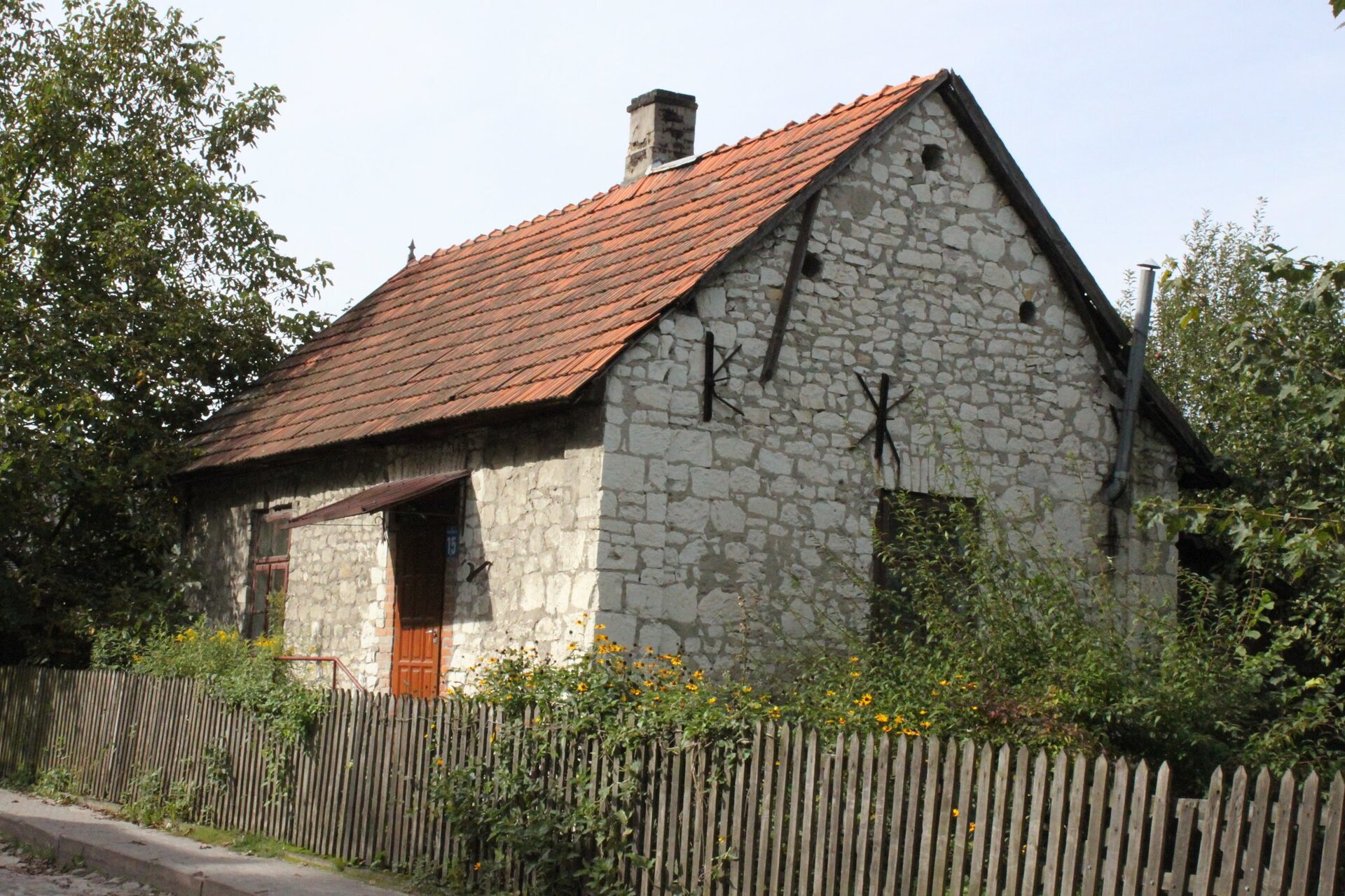 Na fotografii stary dom. Zbudowany jest z wapiennych kamieni. Pokryty czerwoną dachówką. Przed domem znajduje się drewniany płot. Dookoła zielona roślinność. 