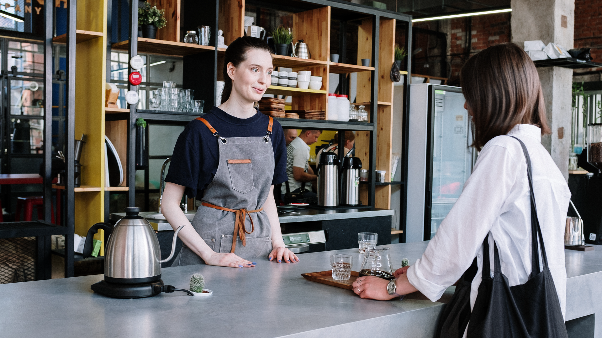 Zdjęcie przedstawia dwie kobiety w barze. Jedna z nich stoi za barem, przodem do kadru. Nosi fartuszek zakładany przez głowę. Druga z kobiet stoi przy barze, tyłem do kadru. Na ramieniu ma torbę. Pomiędzy kobietami na barze stoi taca ze szklankami. Po lewej stronie na barze stoi elektryczny czajnik. W tle widoczne są półki w barze.