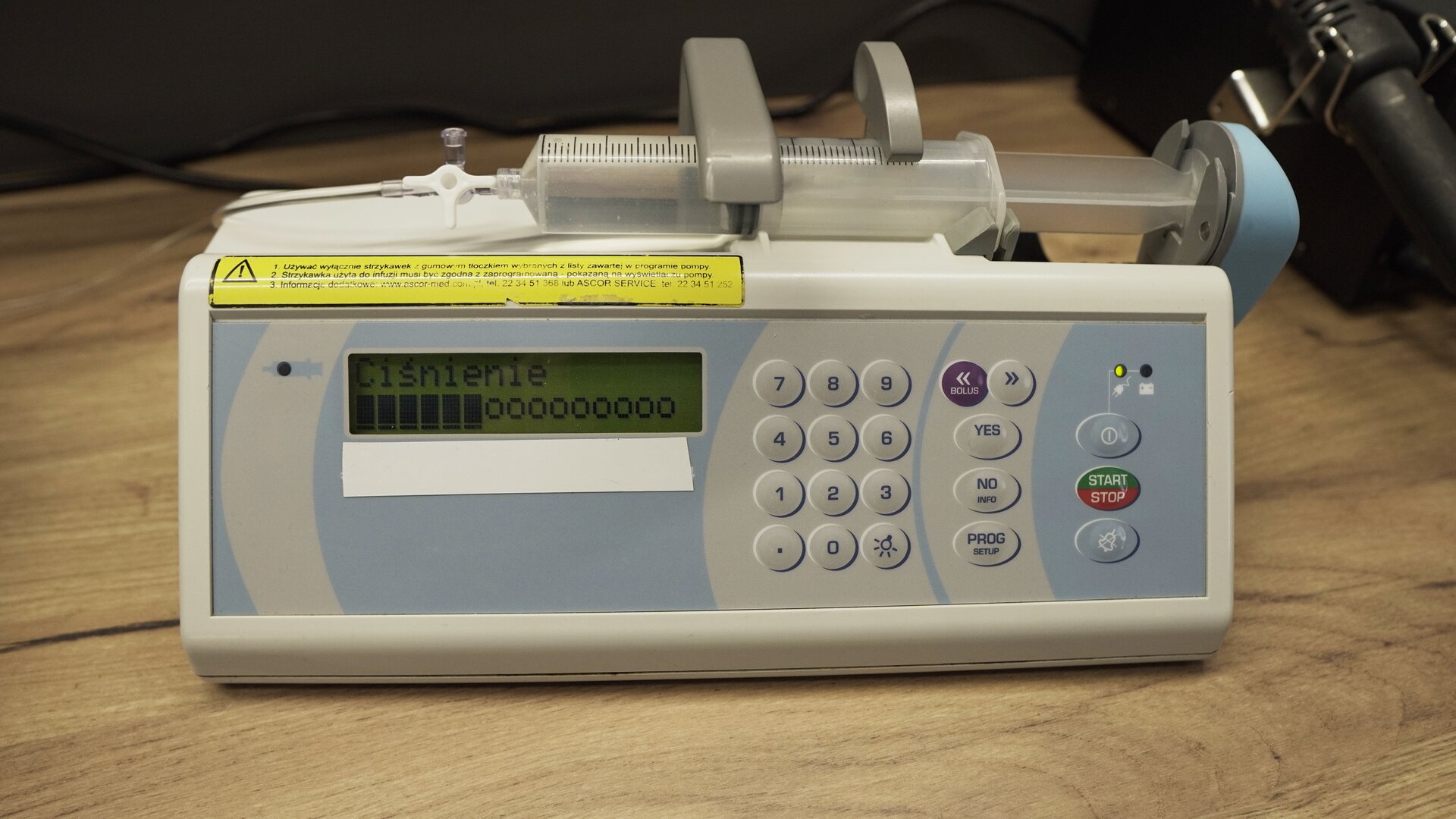 Na zdjęciu strzykawka wypełniona wodą znajduje się w ramieniu pompy infuzyjnej. Na ekranie aparatu wyświetlana jest informacja: "Ciśnienie", a pod spodem od lewej strony 6 wypełnionych prostokątów i dalej 9 okręgów. 