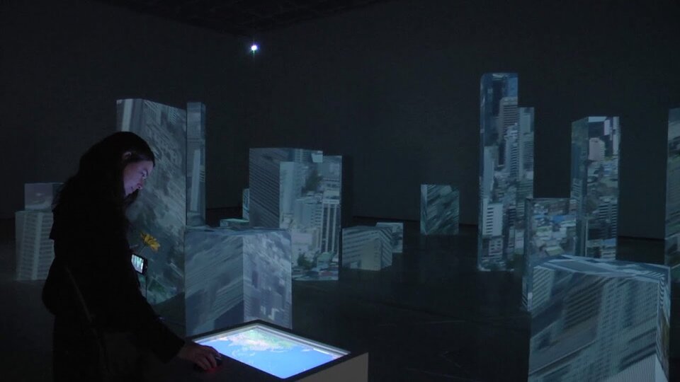 Przedstawiona została kobieta o długich, ciemnych włosach. Ubrana jest w ciemny strój. Stoi przy dużym ekranie zamontowanym w białym pulpicie. Pomieszczenie, w którym się znajduje jest zaciemnione. Rozstawione są w nim statywy w kształcie prostopadłościanów. Wyświetlone zostały na nich fragmenty budowli.