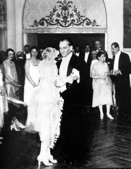 Zdjęcie przedstawia tańczącą parę. Kobieta w białek koronkowej sukni z długimi rękawami i welonem na głowie tańczy ze starszym mężczyzną ubranym w czarny smoking i białą koszulę. Wokół tańczących stoją inni goście, kobiety i mężczyźni ubrani w eleganckie, europejskie stroje. 