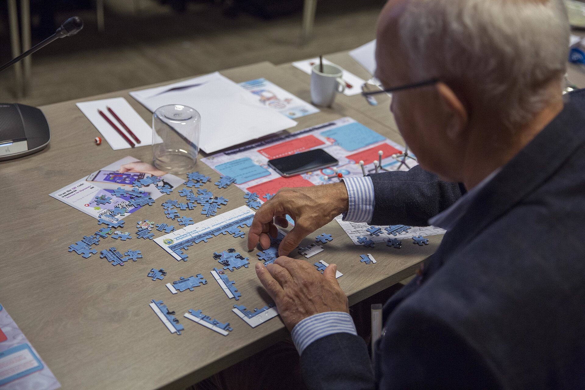  Starszy mężczyzna siedząc przy stole układa puzzle. Na stole obok częściowo ułożonych puzzli znajdują się elementy układanki luźno rozrzucone. Obok stoi słoik odwrócony dnem do góry, filiżanka, w której jest ołówek. Za słoikiem leżą kartki i ołówki, po lewej stronie znajduje się lampka biurowa. Mężczyzna odwrócony jest tyłem. Widać lewy profil i tył głowy. Na twarzy ma okulary korekcyjne. Ubrany jest w garnitur i koszulę w biało- granatowe paski. Mężczyzna pracuje w skupieniu.