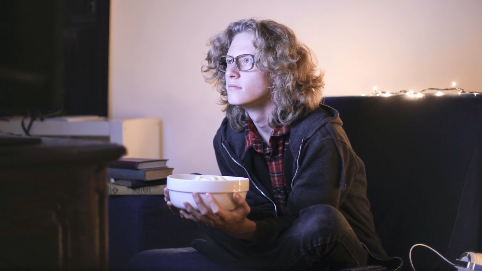 Ilustracja przedstawia młodego mężczyznę w okularach wpatrującego się w ekran telewizora. W ręku trzyma miskę z przekąskami.