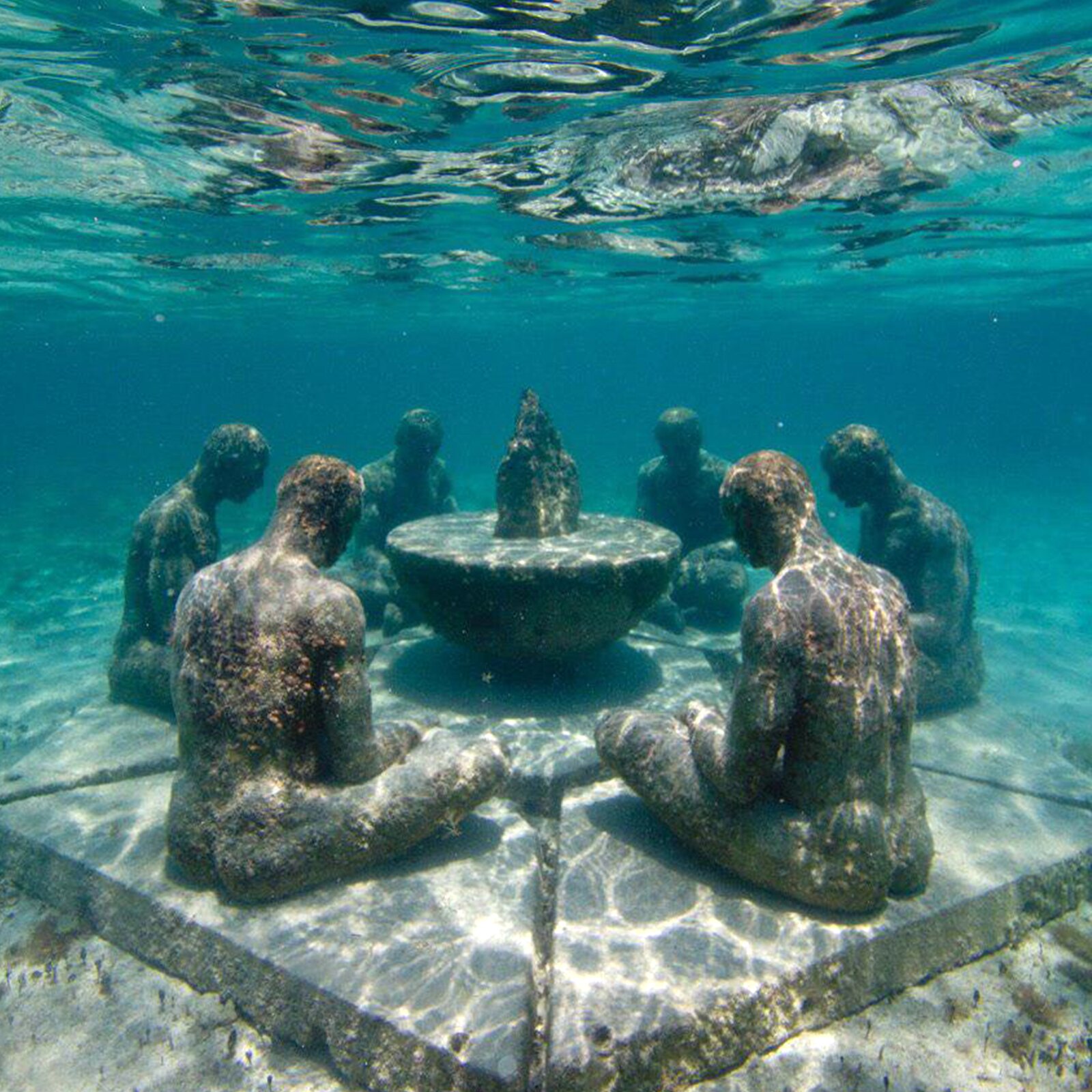 Ilustracja przedstawia sześć rzeźb mężczyzn siedzących na płycie na dnie morza. Są oni zgromadzeni wokół małego stołu, stolika, na którym znajduje się zaostrzony element. Rzeźby sprawiają wrażenie, iż modlą się do tego elementu – ich nogi są skrzyżowane, ręce oparte na udach, a głowy są skierowane w dół.