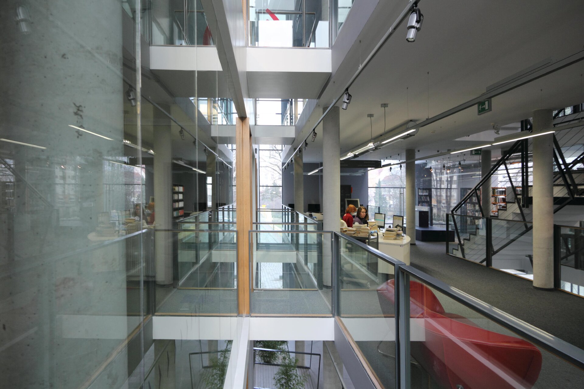 Fotografia ukazuje pomieszczenia biblioteki. Na pierwszym planie znajdują się dwa czerwone fotele stojące za szklaną balustradą. Dalej ukazane są dwie kobiety siedzące przy stanowisku komputerowym. Na biurku znajdują się liczne książki. Po prawej stronie biegną białe schody. Za schodami stoją regały z książkami. Lewa strona zdjęcia to przejście do niewidocznej części budynku oraz błyszcząca ściana, w której odbija się wnętrze pomieszczenia.