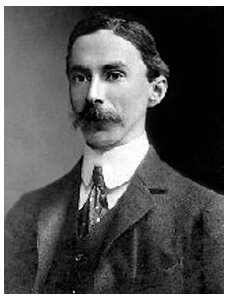 Zdjęcie przedstawia dorosłego eleganckiego mężczyznę o drobnej twarzy i bujnym wąsie. Mężczyzna ma na sobie garnitur.