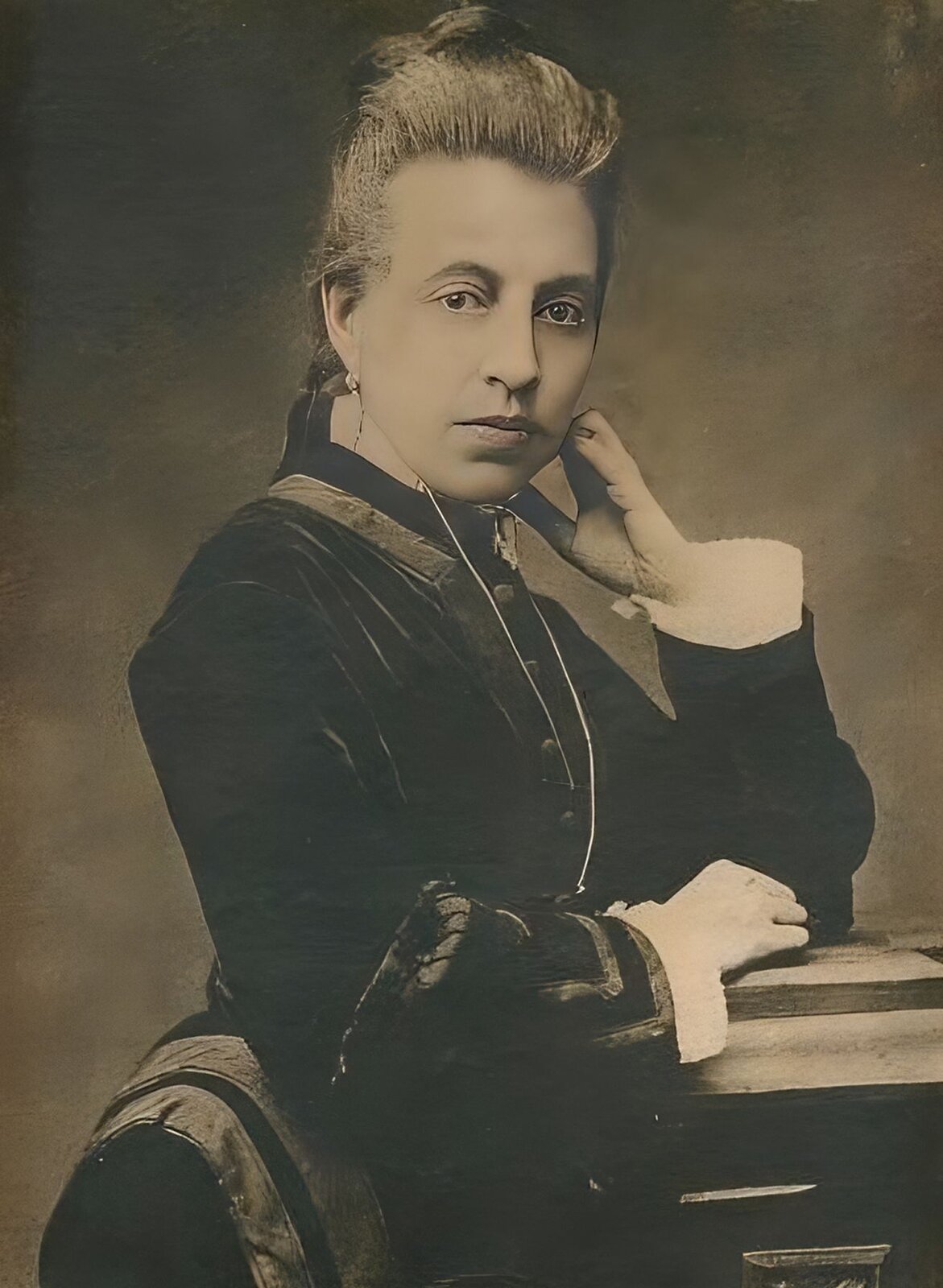 Zdjęcie przedstawia dojrzałą kobietę, która stoi bokiem do obiektywu, ale głowę kieruje wprost do widza. Kobieta ubrana jest w ciemną suknię z długimi rękawami, spod których wystają białe rękawy koszuli. Na szyi zawieszony ma długi wisior na cienkim łańcuszku. Włosy ma upięte w wysokiego koka. Kobieta prawą dłonią podpiera głowę, a lewą położyła na stole. Pozuje na jednolitym tle. Zdjęcie jest czarno‑białe.