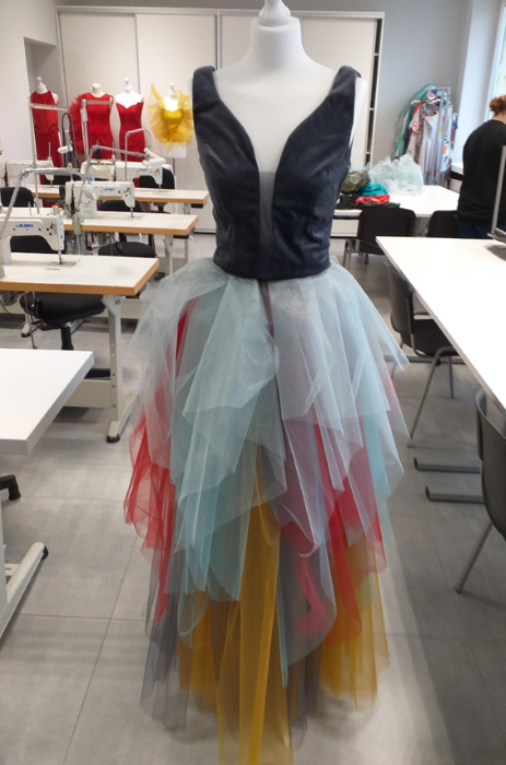 Zdjęcie przedstawia wiszącą na manekinie sukienkę. Ma czarny wąski gorset oraz tiulową, lejącą i wielowarstwową spódnicę złożoną z materiałów w kolorach turkusowym, żółtym i czerwonym.