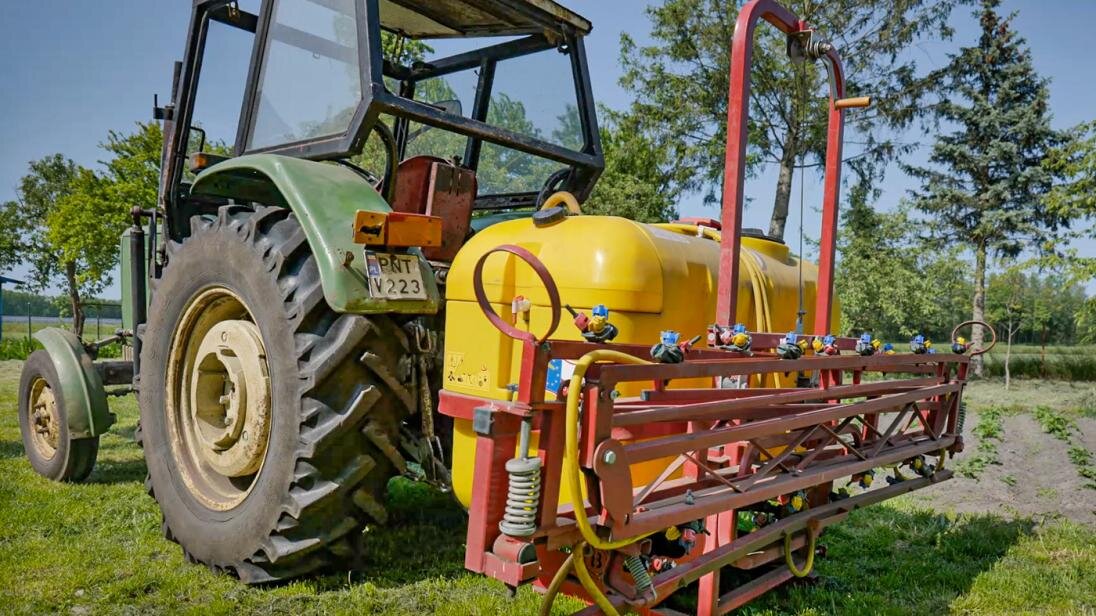 Zdjęcie opryskiwacza zapiętego do ciągnika. Urządzenie składa się z dużego zbiornika oraz systemu przewodów rurowych do rozpylania cieczy.
Nagranie tożsame z treścią opisu.