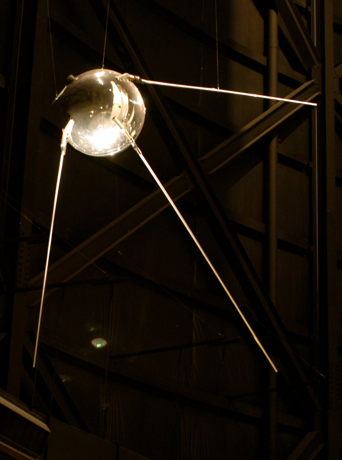Zdjęcie przedstawia pierwszego sztucznego satelitę Ziemi – SPUTNIK 1. Tło ciemne, prawie czarne. Na zdjęciu widoczne urządzenie przypominające dużą, srebrną kulę z trzema cienkimi „nóżkami” odchodzącymi od niej. Powierzchnia błyszcząca.