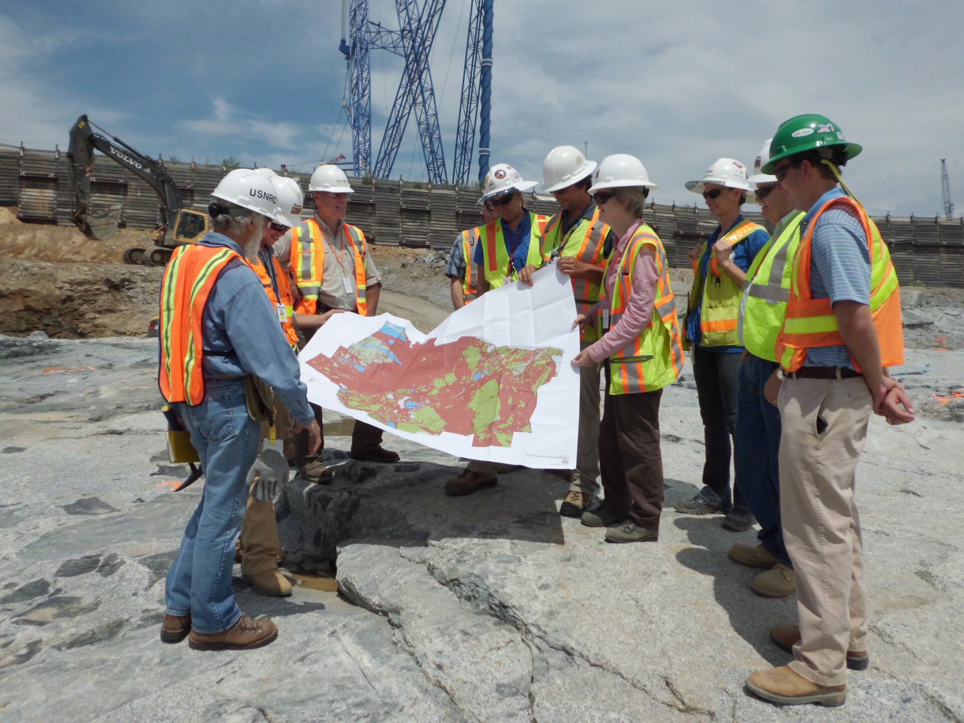 Ilustracja ukazuje teren budowy, na nim grupa budowlańców, część z nich trzyma wielką mapę analogową.