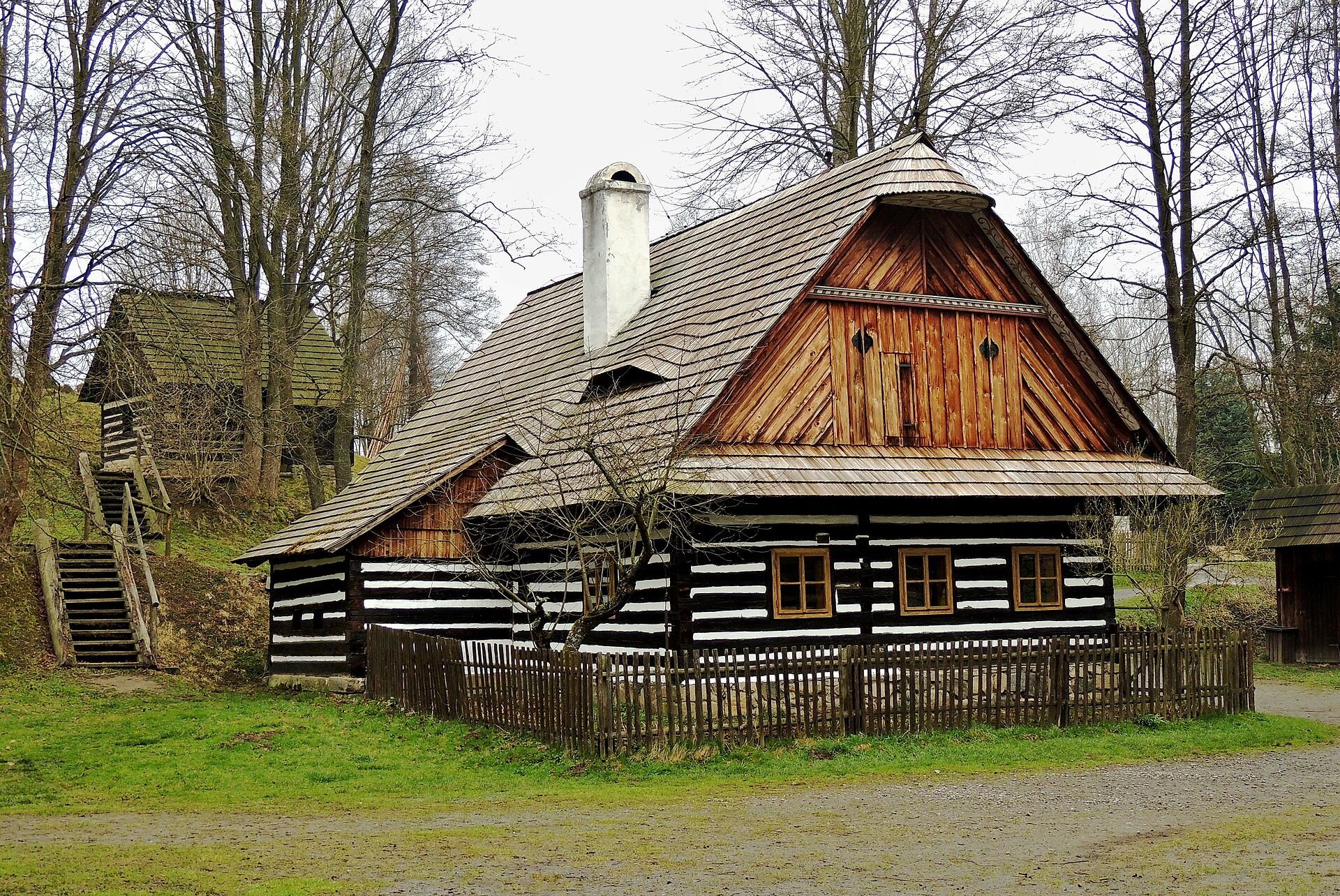 Fotografia drewnianej chałupy położonej na terenie skansenu. Dookoła budynku drewniany płot, a dalej droga. W tle inne budynki.