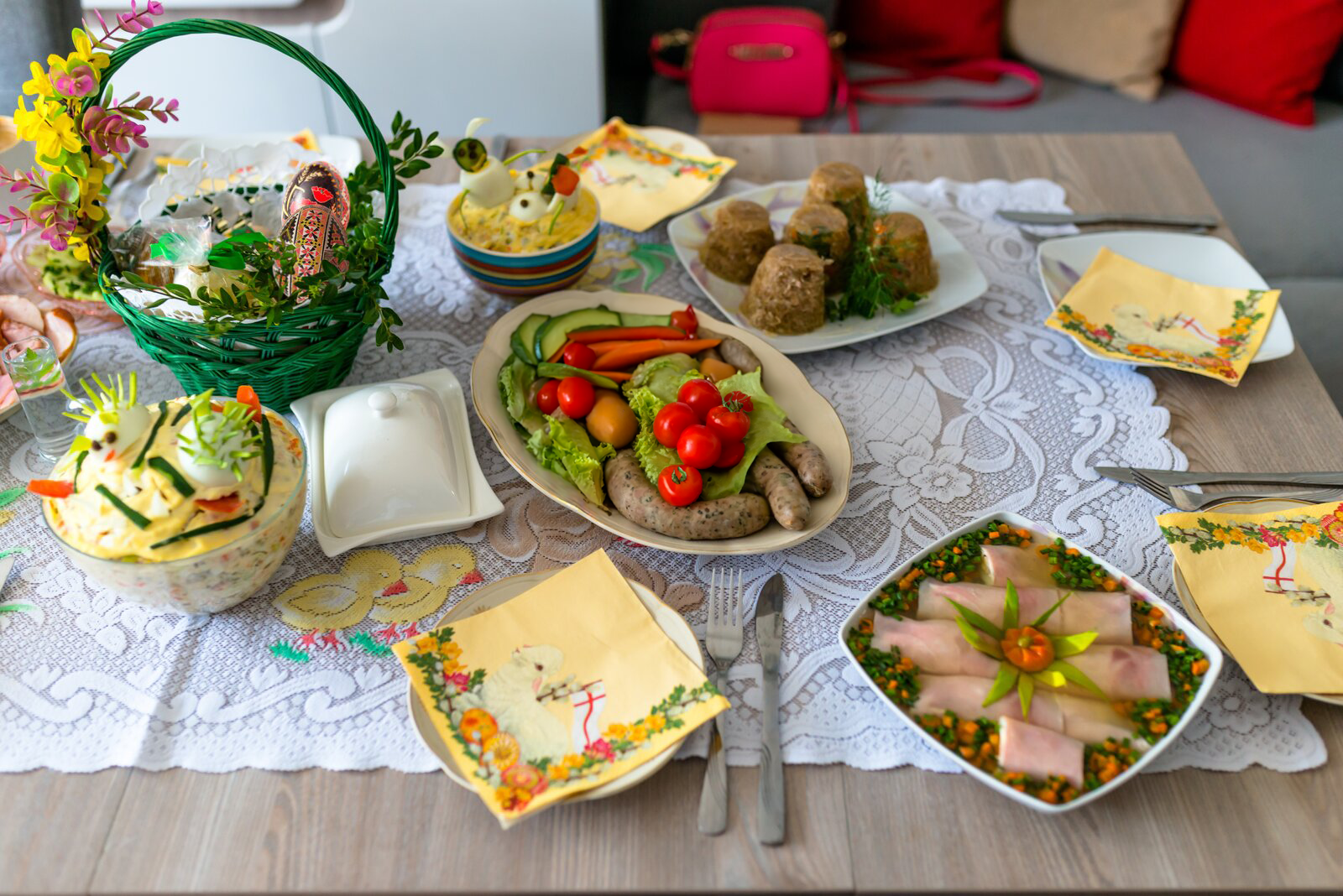 Zdjęcie przedstawia stół wielkanocny. Na dużej białej serwecie ustawiono różne potrawy: kiełbasy, galaretki wieprzowe, szynkę w galarecie, sałatki warzywne. Po lewej stronie stoi zielony koszyk ze święconką, a obok niego zamknięta maselniczka. Dookoła potraw znajdują się cztery talerzyki wraz ze sztućcami. Na każdym talerzyku leży kwadratowa papierowa serwetka z motywem wielkanocnym.