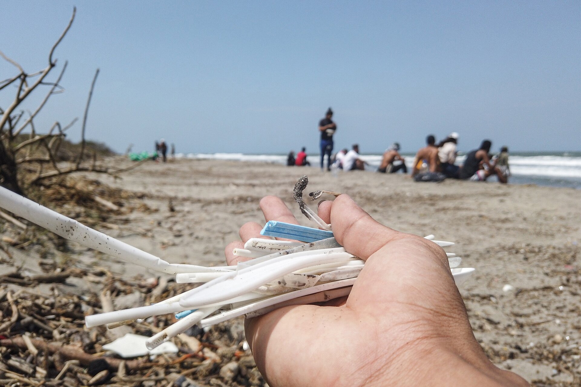 Na pierwszym planie dłoń na której leżą kawałki plastiku. W tle plaża i grupa ludzi siedzących nad morzem.