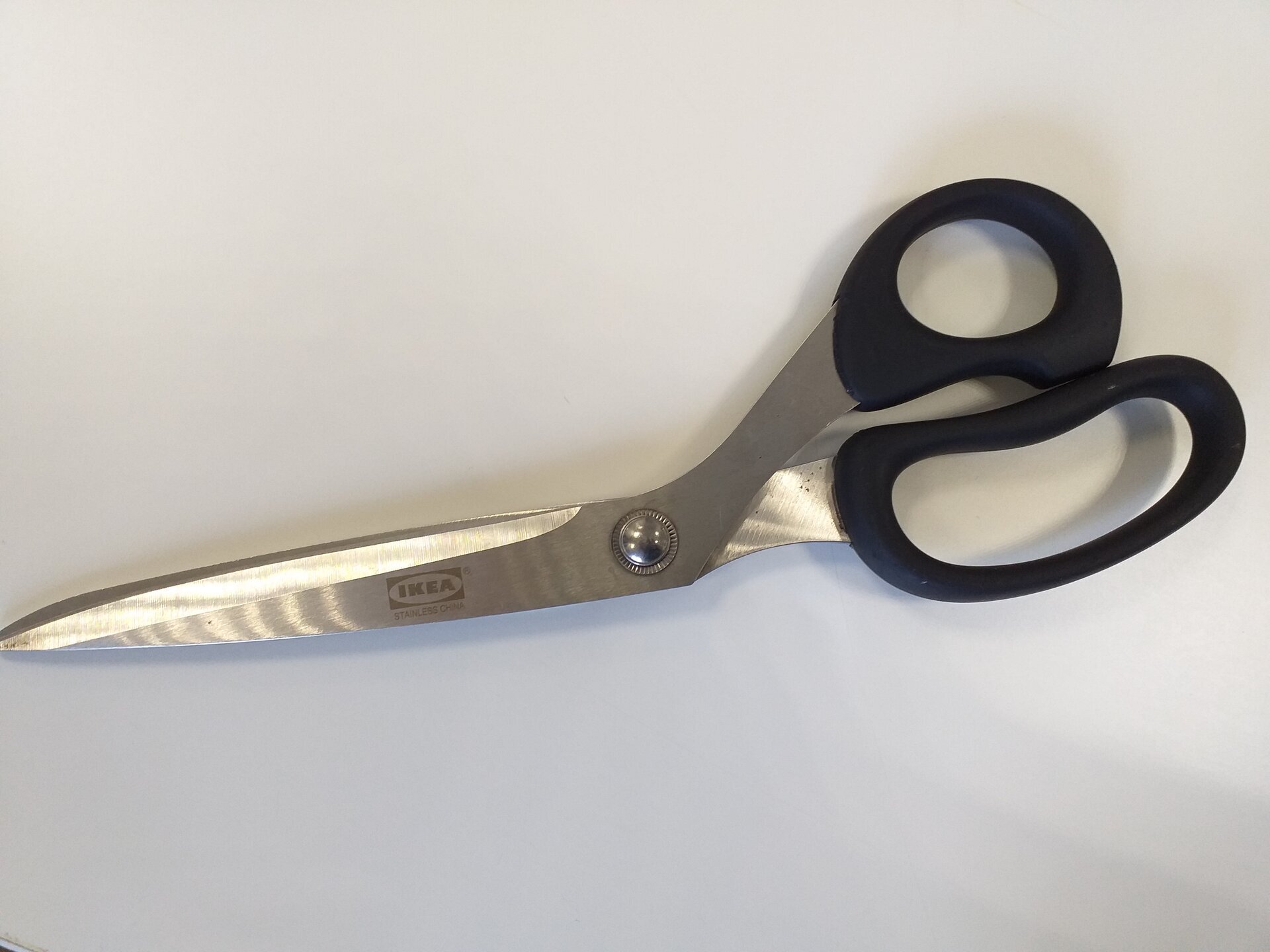 Zdjęcie przedstawia nożyce krawieckie. Składają się one z długich, metalowych ostrzy oraz z plastikowych rączek dopasowanych do kształtu dłoni i kciuka.