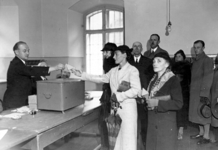 Zdjęcie przedstawia wnętrze sali. Z lewej strony ustawione są stoły, za którymi siedzi dwóch mężczyzn, jeden stoi i odbiera od kobiety kopertę. Kobieta stoi wśród innych osób ustawionych w kolejce przed stołami.