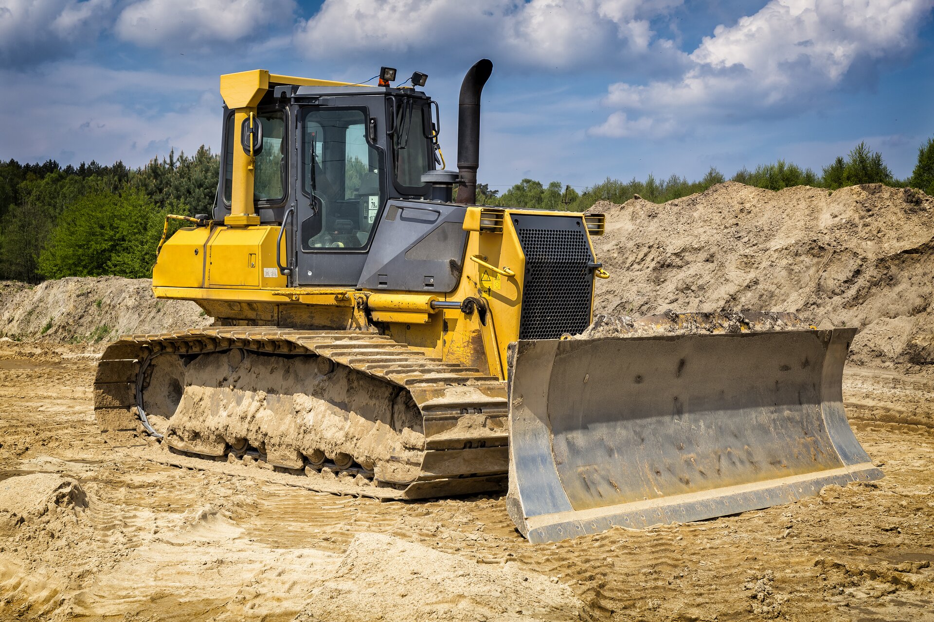 Zdjęcie przedstawia spycharkę. Widoczna jest maszyna w ujęciu z boku, stojąca na rozjeżdżonej ziemi. W przedniej części widoczne  jest ramię spycharki z szerokim lemieszem, czyli elementem pracującym. W tyle widoczna jest kabina operatora spycharki. W tle widoczne są hałdy ziemi.

Opisy obiektów prezentowanych na grafikach są załączone w treści galerii zdjęć.