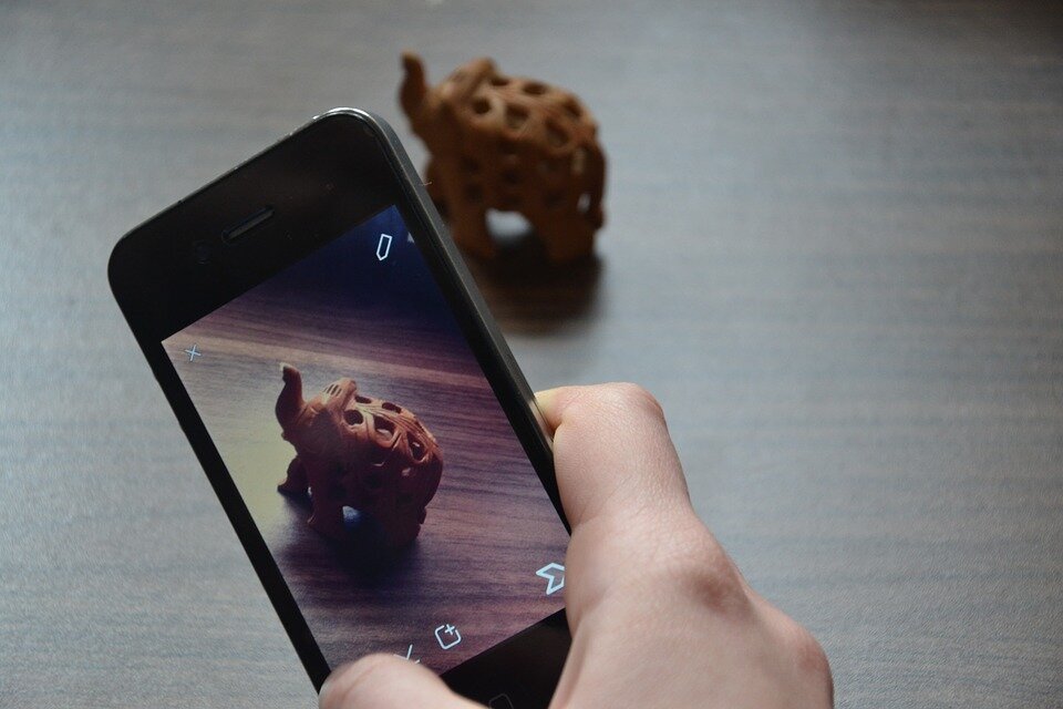 Fotografia barwna przedstawiająca trzymany przez kogoś w dłoni smartfon. W tle widać drewnianą figurkę słonia - na ekranie smartfona widać edytor zdjęć. Ktoś właśnie zrobił zdjęcie figurki.