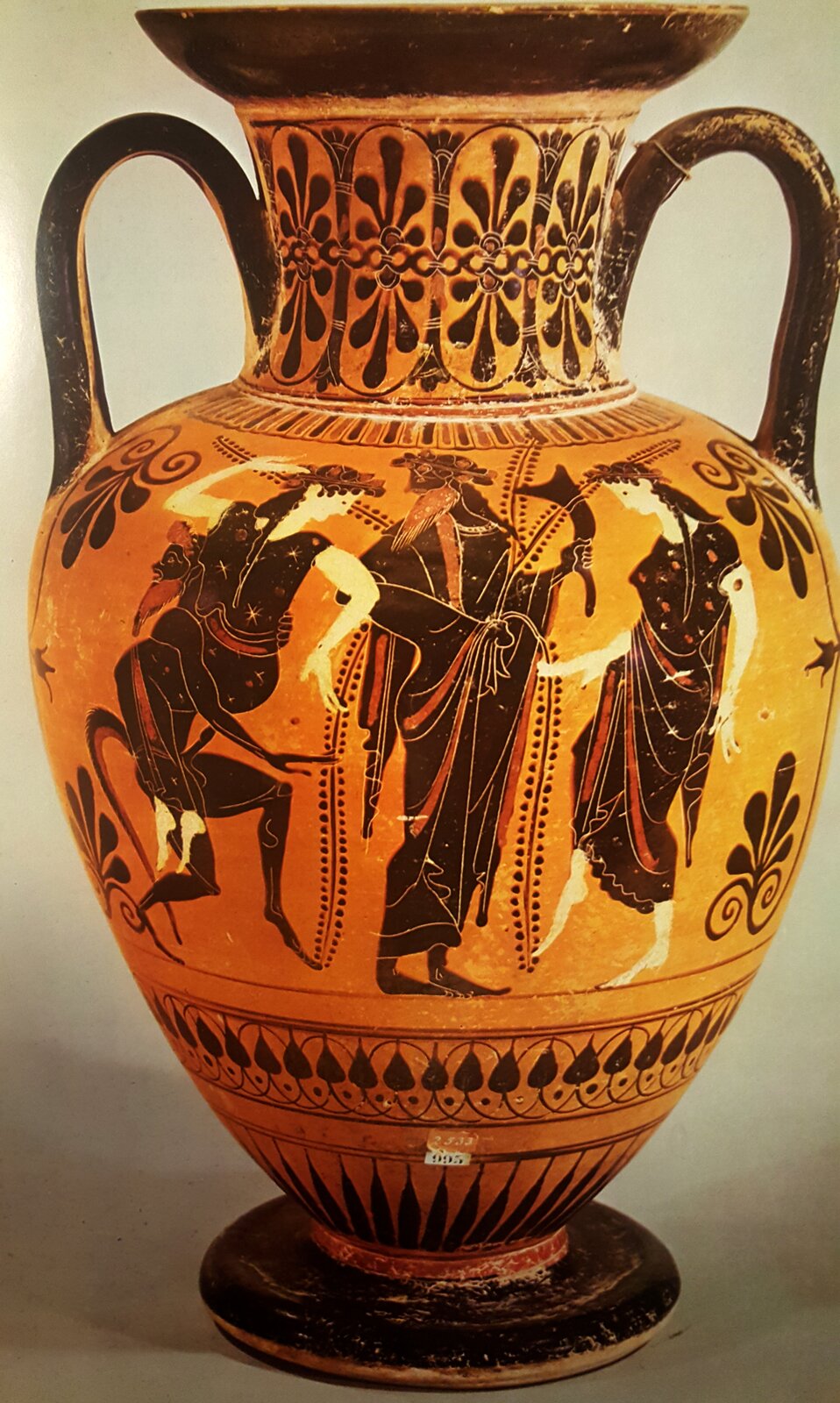 Ilustracja przedstawia starożytną wazę w stylu czarnofigurowym z dwoma czarnymi uszami. Pośrodku znajdują się postacie: stojący mężczyzna i tańczące po obu stronach kobiety. Dolna i górna część są dekorowane motywami kwiatowymi oraz wypełnione czarną barwą. Tło jest czerwonawo-pomarańczowe.