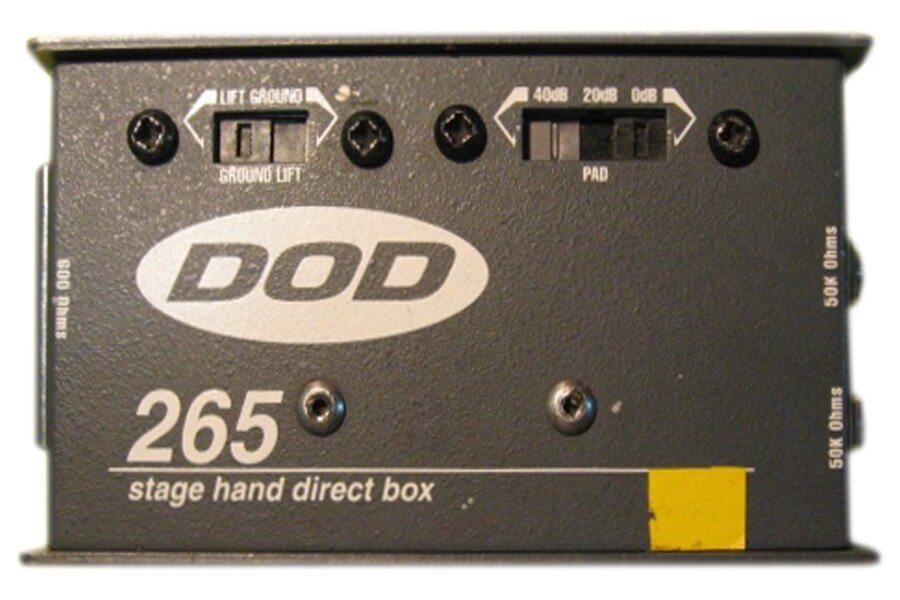 Zdjęcie przedstawia urządzenie d‑box, które ma kształt szarego prostopadłościanu w ujęciu frontalnym. Na urządzeniu, w górnej części widoczne są dwa prostokątne suwaki. Po prawej i lewej stronie suwaków widoczne są pojedyncze czarne kółka z wpisanym w nie czarnym krzyżykiem – główki śrubek. Poniżej i powyżej suwaków widoczne są białe napisy. Poniżej po lewej stronie widoczna jest biała elipsa, w którą wpisany jest szary napis DOD. Poniżej napisu, po lewej stronie, widoczne są białe cyfry 256, podkreślone białą poziomą linią, poniżej której napis stage hand direct box. Po prawej stronie, poniżej białej poziomej linii, widoczny jest żółty kwadrat.