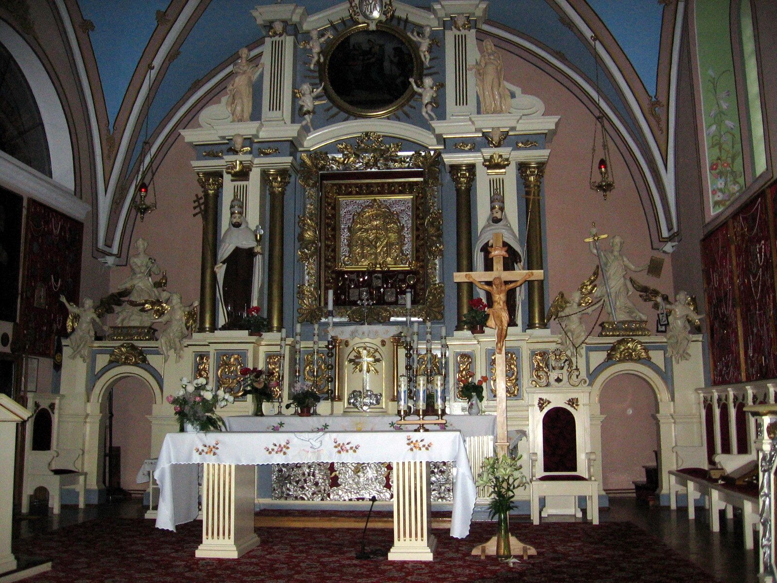 Zdjęcie przedstawia ołtarz w kościele. W centralnej jego części znajduje się złoty obraz, wokół którego stoją rzeźby ludzkie. Pod obrazem znajduje się złoty krzyż. Przed ołtarzem stoi stół nakryty białym obrusem. Po jego prawej stronie stoi drewniany krzyż. Sufit kościoła jest niebieski.