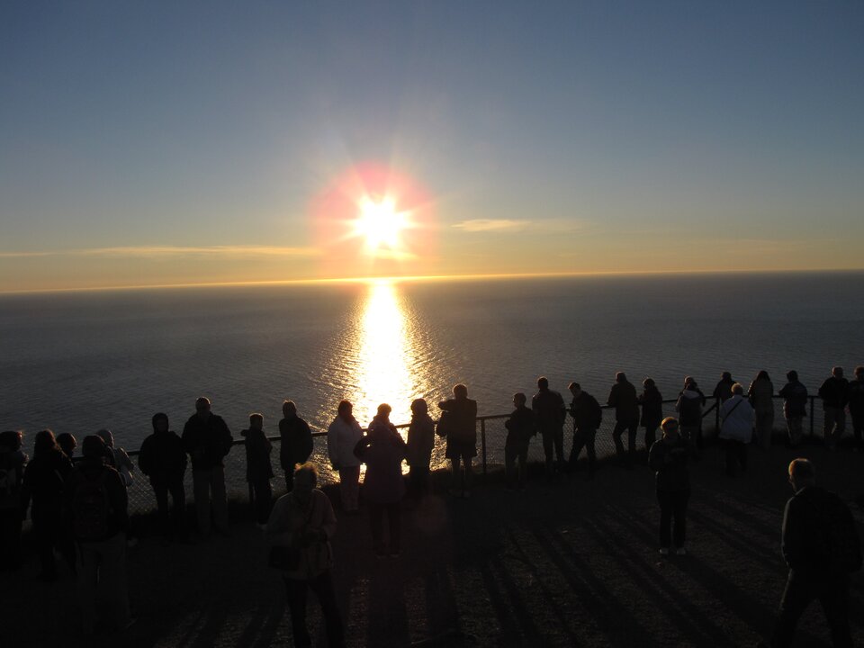 Zdjęcie przedstawia słońce znajdujące się nieco powyżej horyzontu nad morzem. Na brzegu, przy barierce stoją jeden obok drugiego ludzie, którzy obserwują słońce.